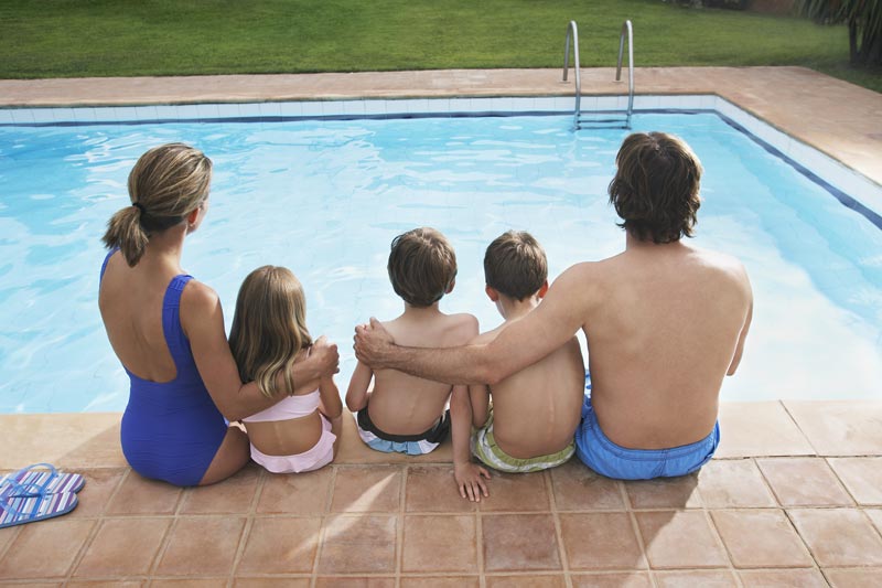 family with young kids sitting on edge of outdoor inground pool