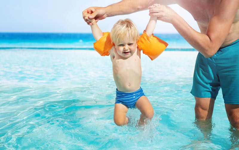 father dipping toddler boy into water with arm floats on
