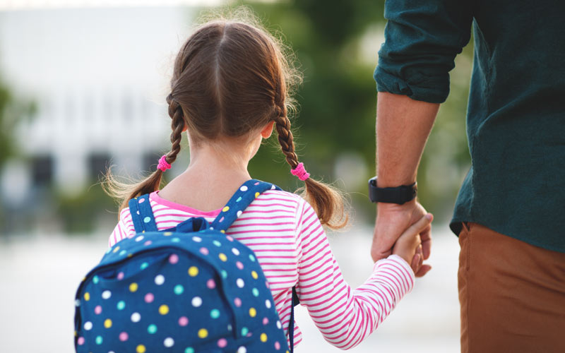 father holding young daughters hand as she goes with backpack on to school