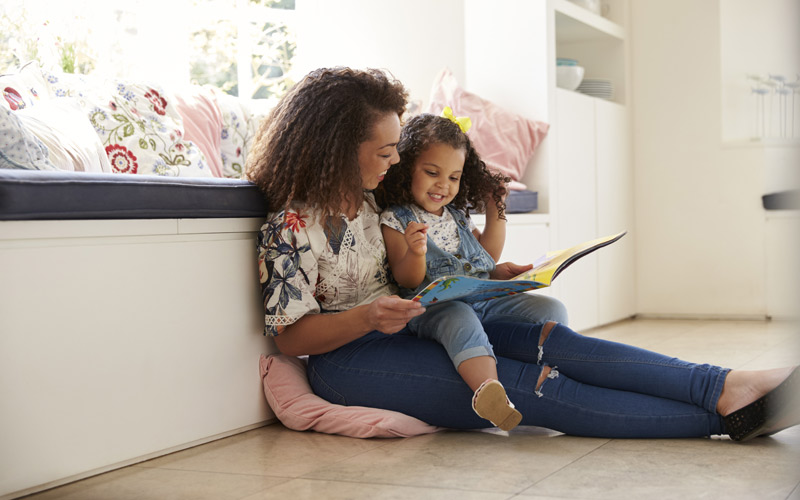 mom sitting on floor reading book to young daughter