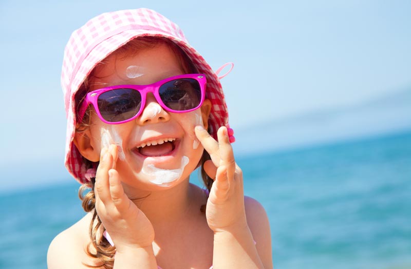 smiling little girl wearing sunglasses and sunhat applying sunblock lotion