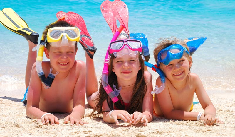 three siblings wearing scuba gear lying on the beach by the ocean