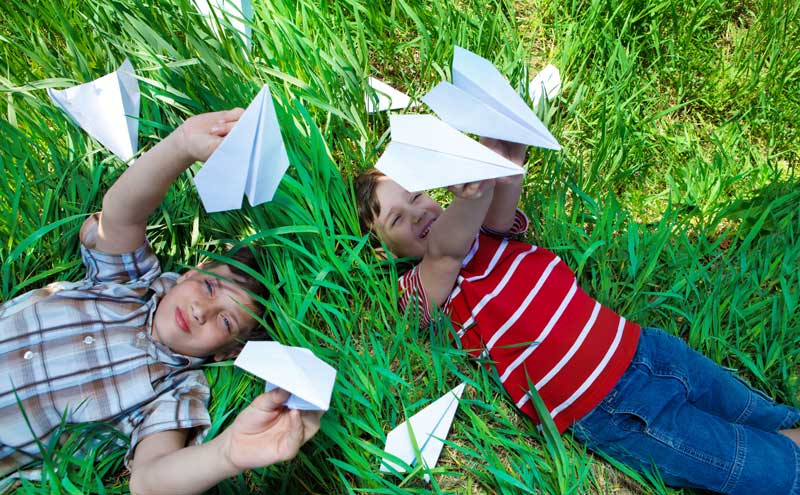 two boys lying on the grass and in the shade while playing with paper airplanes
