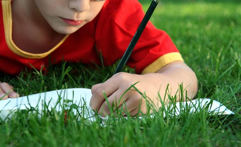 young boy writing in a book while lying in the green grass