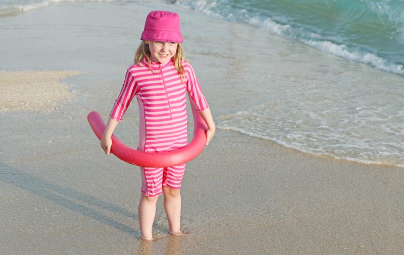 young girl standing in sand by waves wearing sun protective clothing